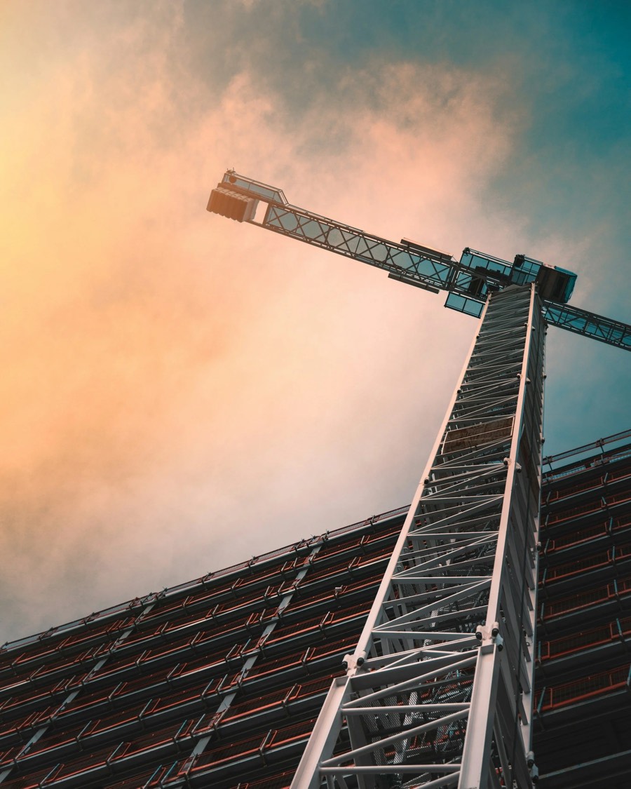 A close-up view of a modern construction crane with a building under construction in the background, taken during sunset with colorful sky.