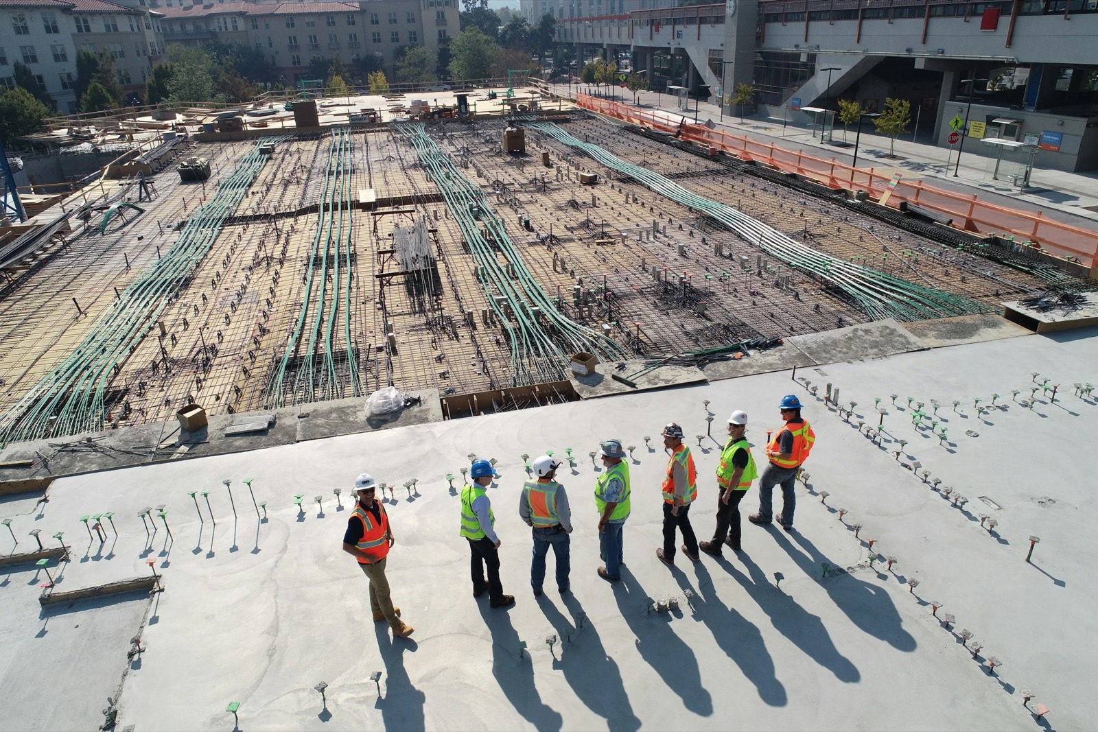 Aerial view of seven construction workers in high-visibility vests and hard hats standing on a concrete slab beside reinforcement and conduit.