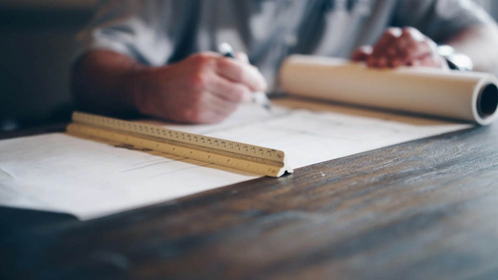 A person is sitting at a wooden desk, writing on a piece of paper with a pen. A yellow ruler and a rolled-up sheet of paper are on the desk.